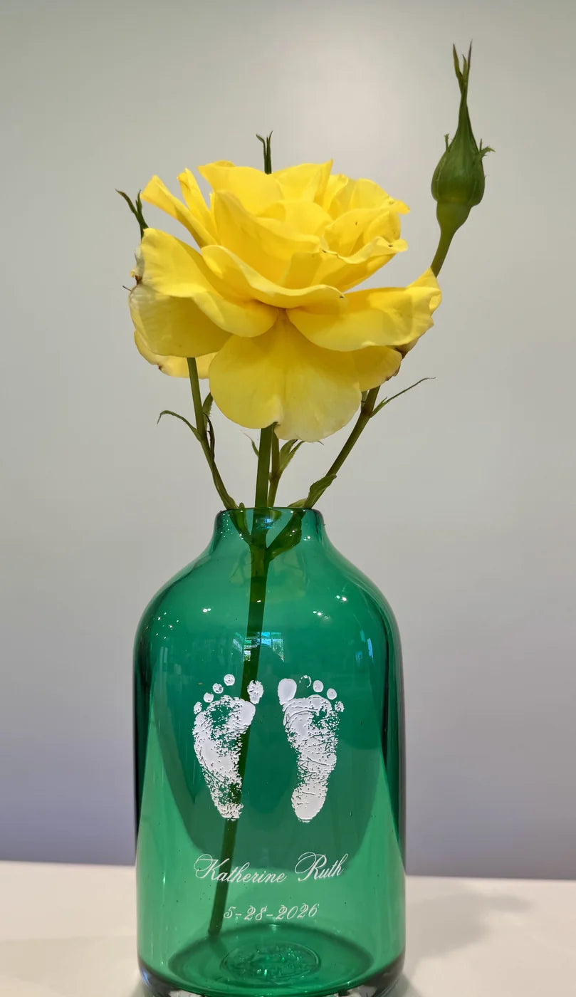 A green glass keepsake vase engraved with baby footprints, holding a single yellow rose