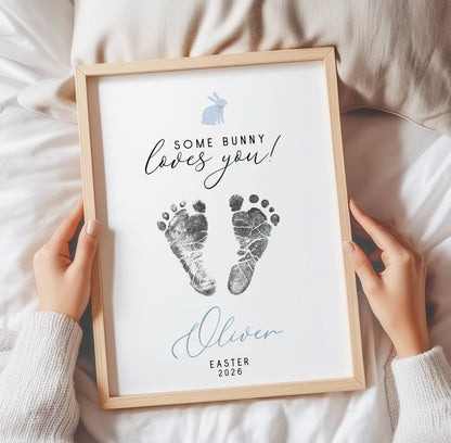 Framed keepsake with baby footprints and text 'Some bunny loves you! Oliver Easter 2026' held by hands on a soft surface.