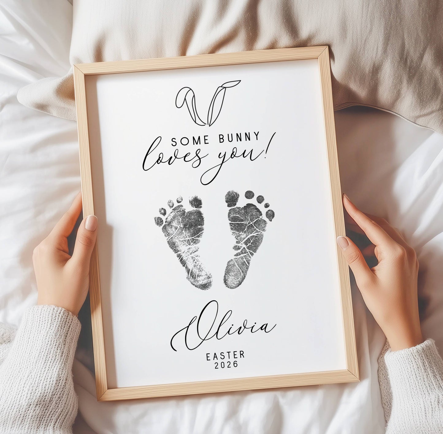 Framed keepsake with baby footprints and text 'Some bunny loves you! Olivia Easter 2026' held by hands.