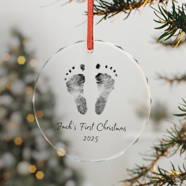 Custom glass ornament with footprints, First Christmas 2025' text, hanging on a tree branch.