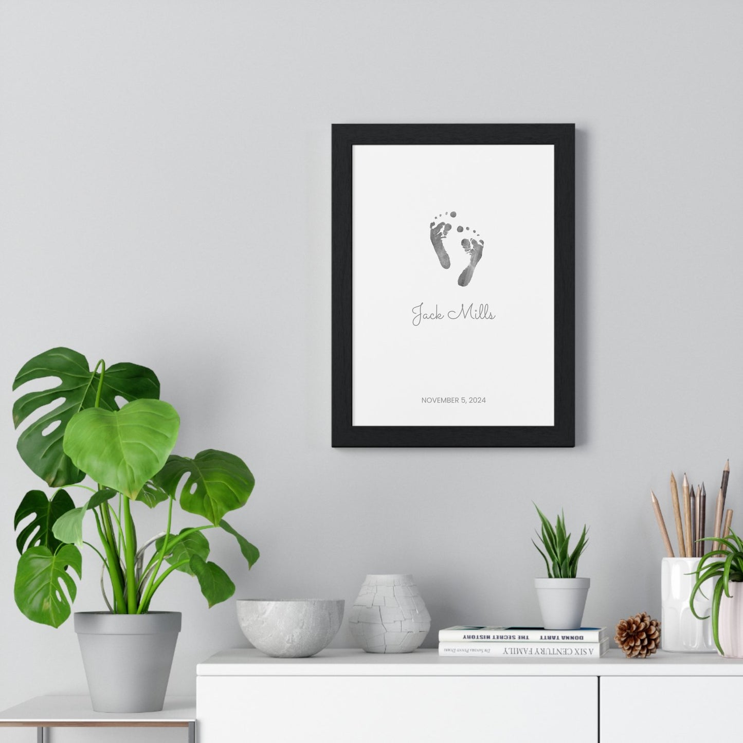 Framed keepsake print with baby footprints on a wall above a white cabinet with decorative items.