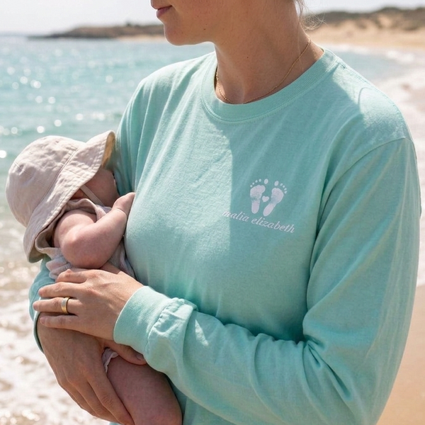 Woman in a light blue shirt with a baby's footprints holding a baby on a beach