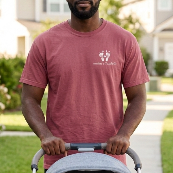 Man wearing a footprint t-shirt pushing a stroller with a baby inside on a sidewalk in a residential area.
